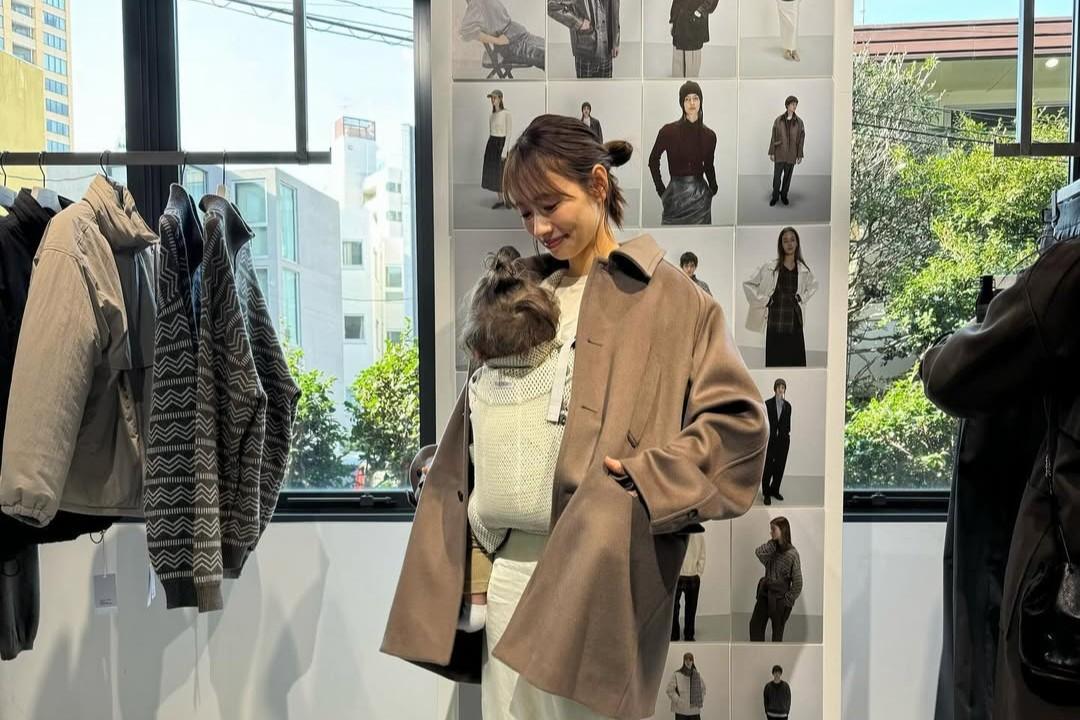 A woman in a store smiles while holding a small dog inside her coat. Clothing is displayed on racks and a wall in the background.