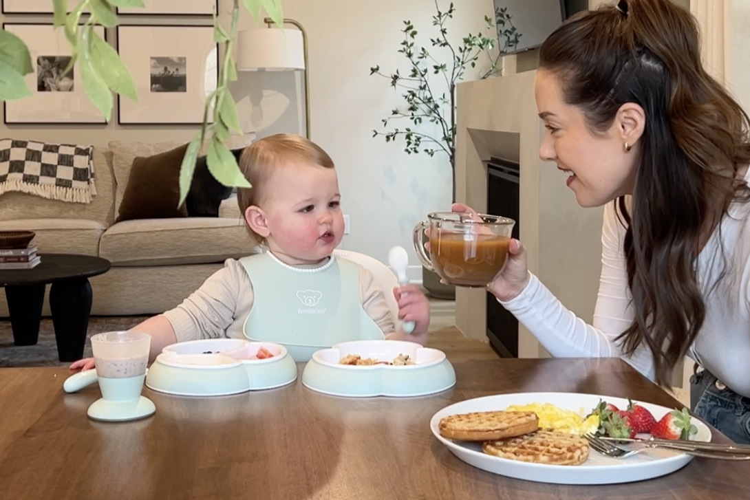 A woman smiles at a baby seated in a high chair with a meal. The table has food, including waffles, eggs, and strawberries. A cozy living room is in the background.