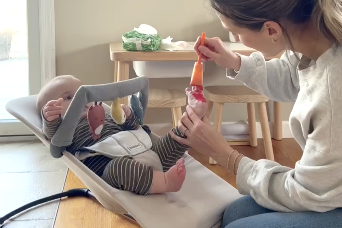 A woman paints a baby's foot while the baby sits in a bouncer, while toys hang from the seat's arch.