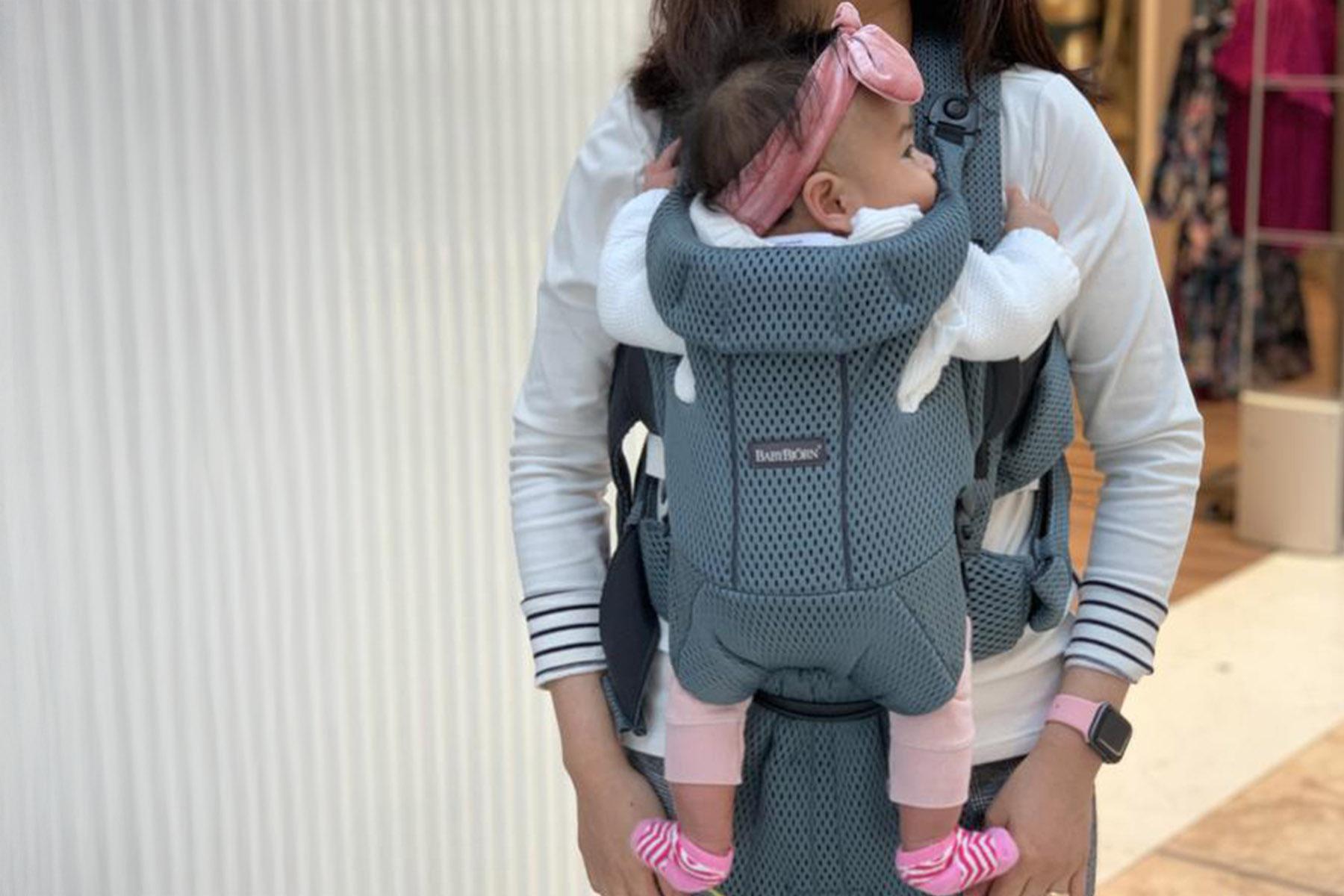Person carrying infant in gray mesh front carrier; baby wearing pink headband, white top, pink leggings and striped socks.