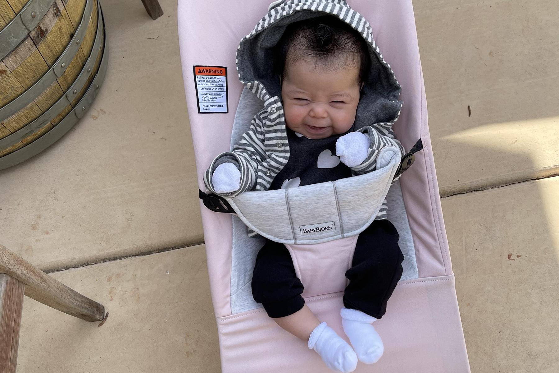 A smiling baby in a striped hoodie sits in a pink baby bouncer, with a wooden barrel and chair nearby on a patio.