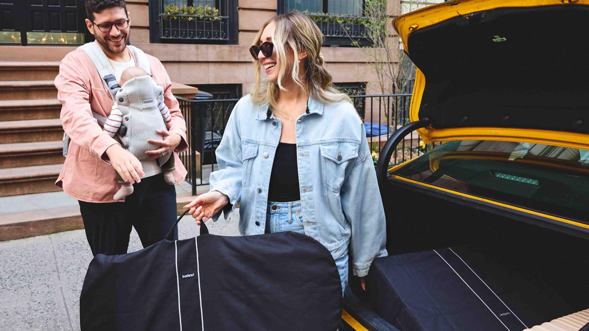 A smiling couple loads luggage into a car trunk. The man holds a baby in a carrier, and the woman wears sunglasses and a denim jacket.
