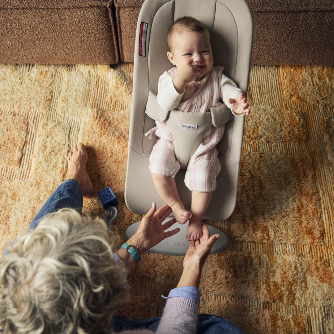 A smiling baby in a bouncer with a person playfully holding the baby's feet on a patterned carpet.