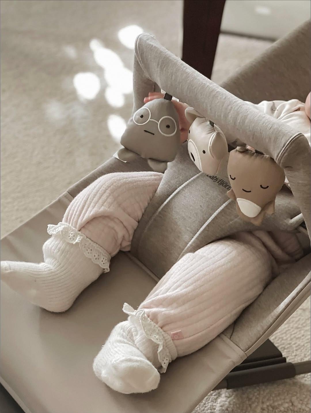 Baby in a bouncer with soft pink pants and white socks, surrounded by plush toys.