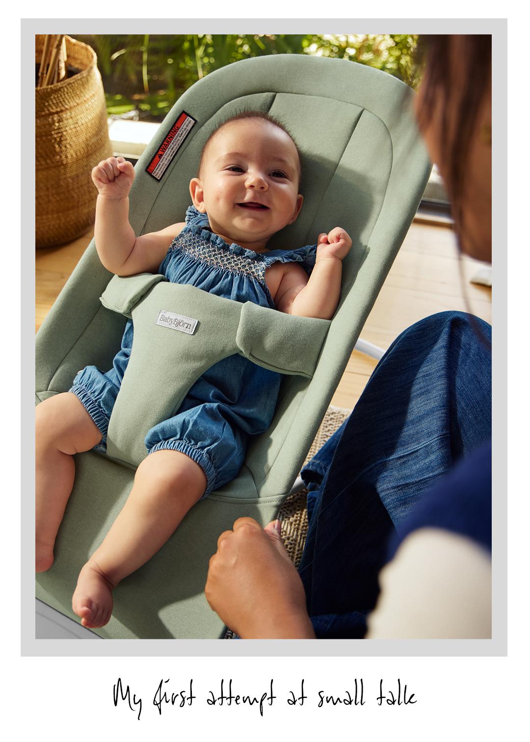 A smiling baby in a blue romper sits in a green bouncer. A person is interacting with the baby, creating a warm, playful scene.