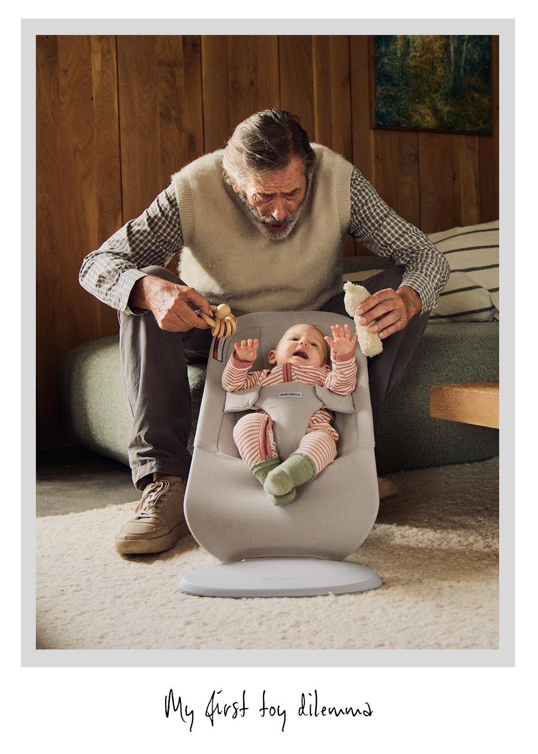 An older man offering a baby, seated in a bouncer, two toys: a plush toy and a wooden rattle, in a cozy room setting.