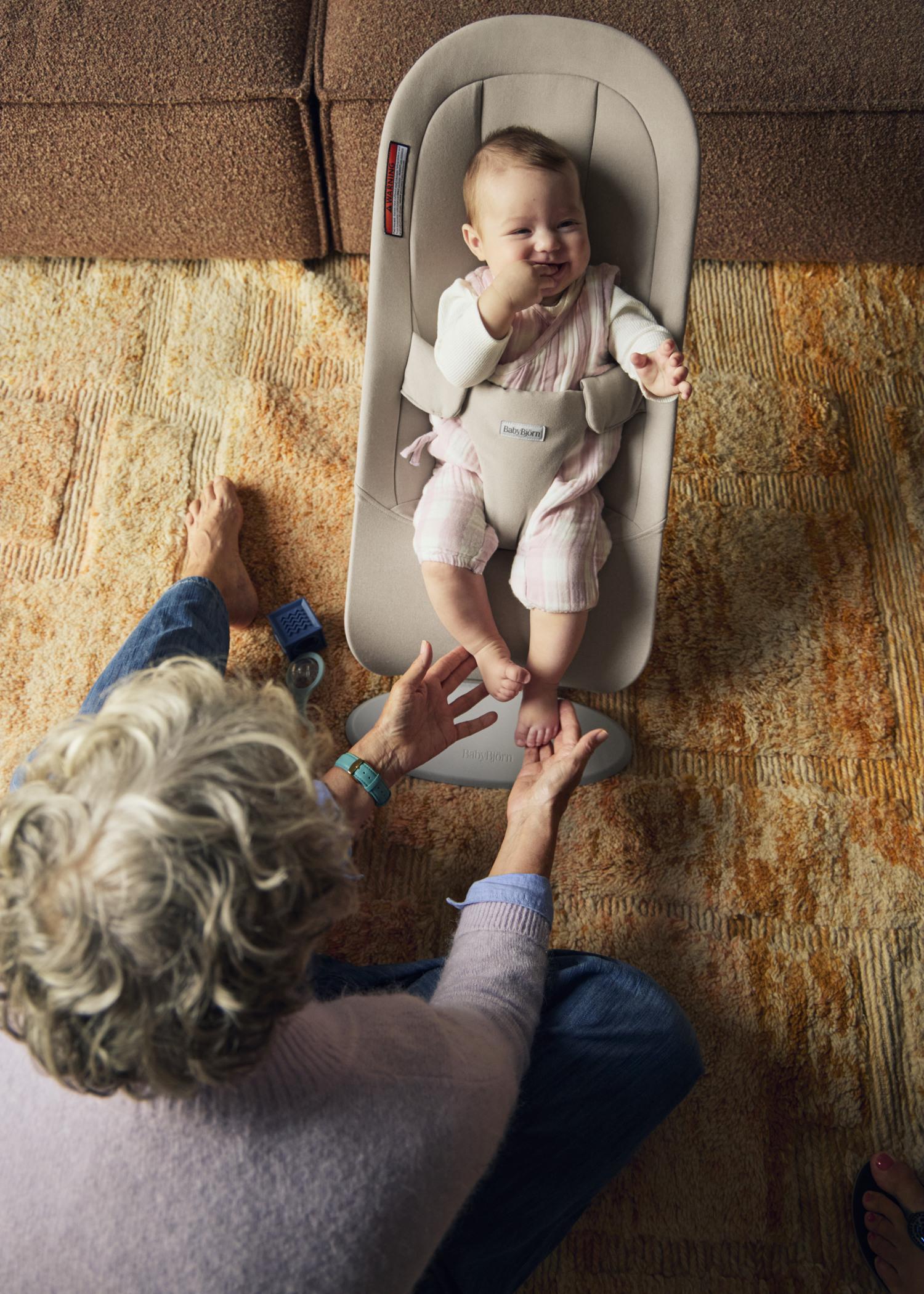 A smiling baby in a bouncer is being gently played with by an adult sitting on a patterned carpet.
