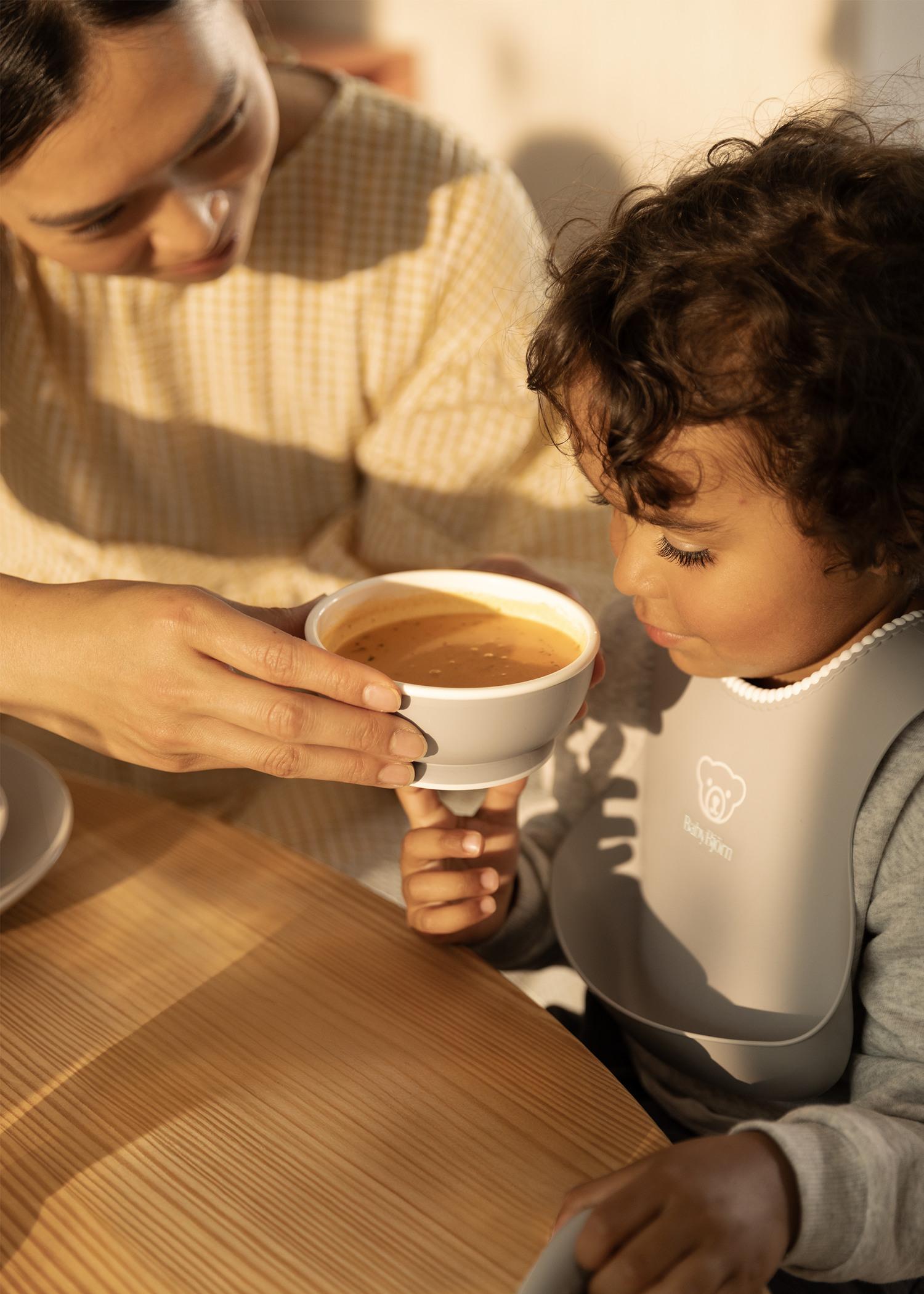 A woman helps a child drink from a cup while sitting at a wooden table. The child wears a bib and looks at the cup attentively.