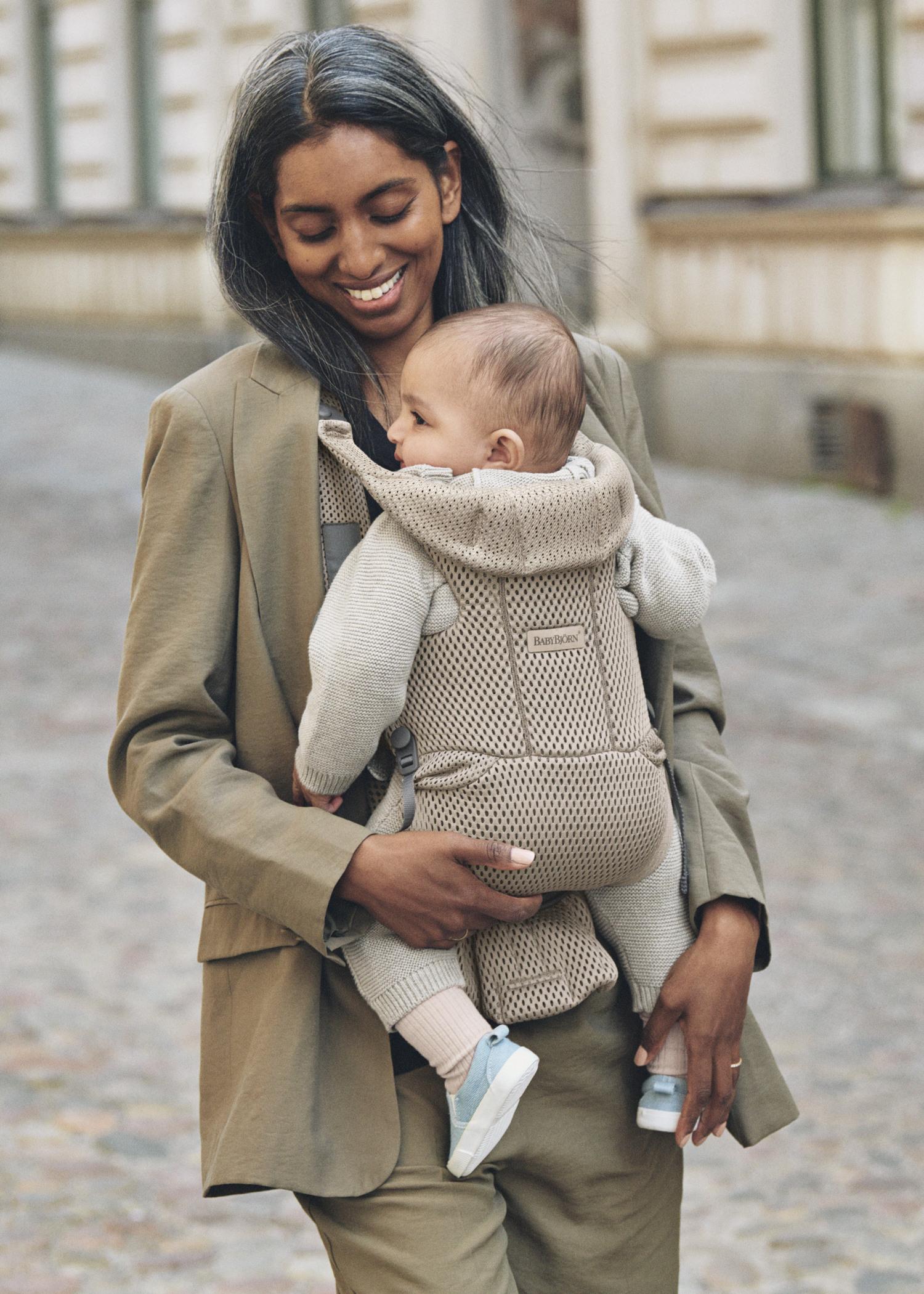 A smiling woman walks down a cobblestone street, carrying a baby in a beige carrier. Both are dressed in neutral tones.