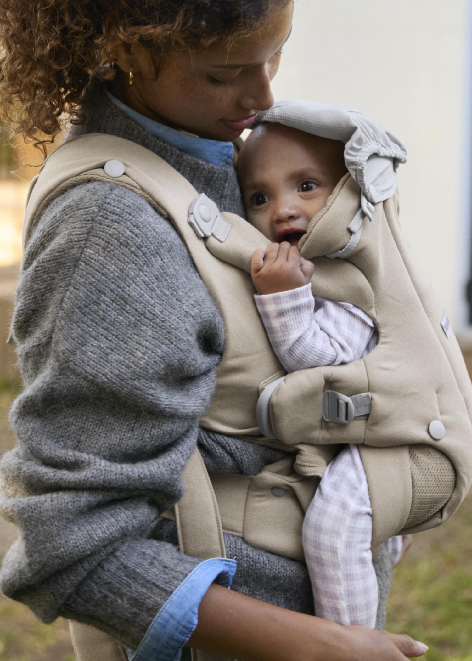 A woman wearing a gray sweater holds a baby in a beige baby carrier. The baby, wearing pink outfit, looks curious and comfortable.