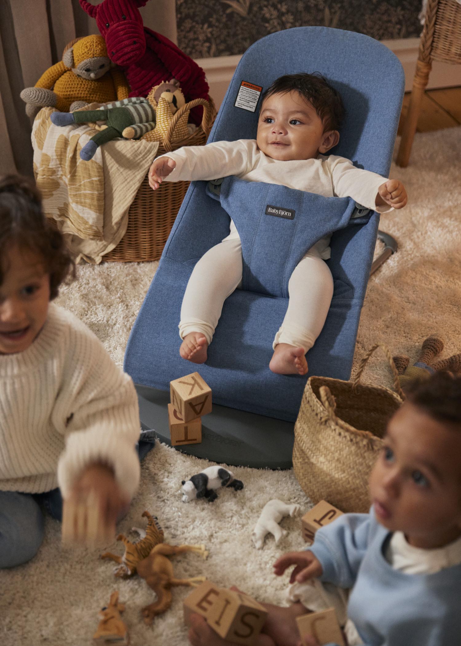 Baby in a blue bouncer, surrounded by two toddlers playing with wooden blocks and toy animals on a soft carpet.