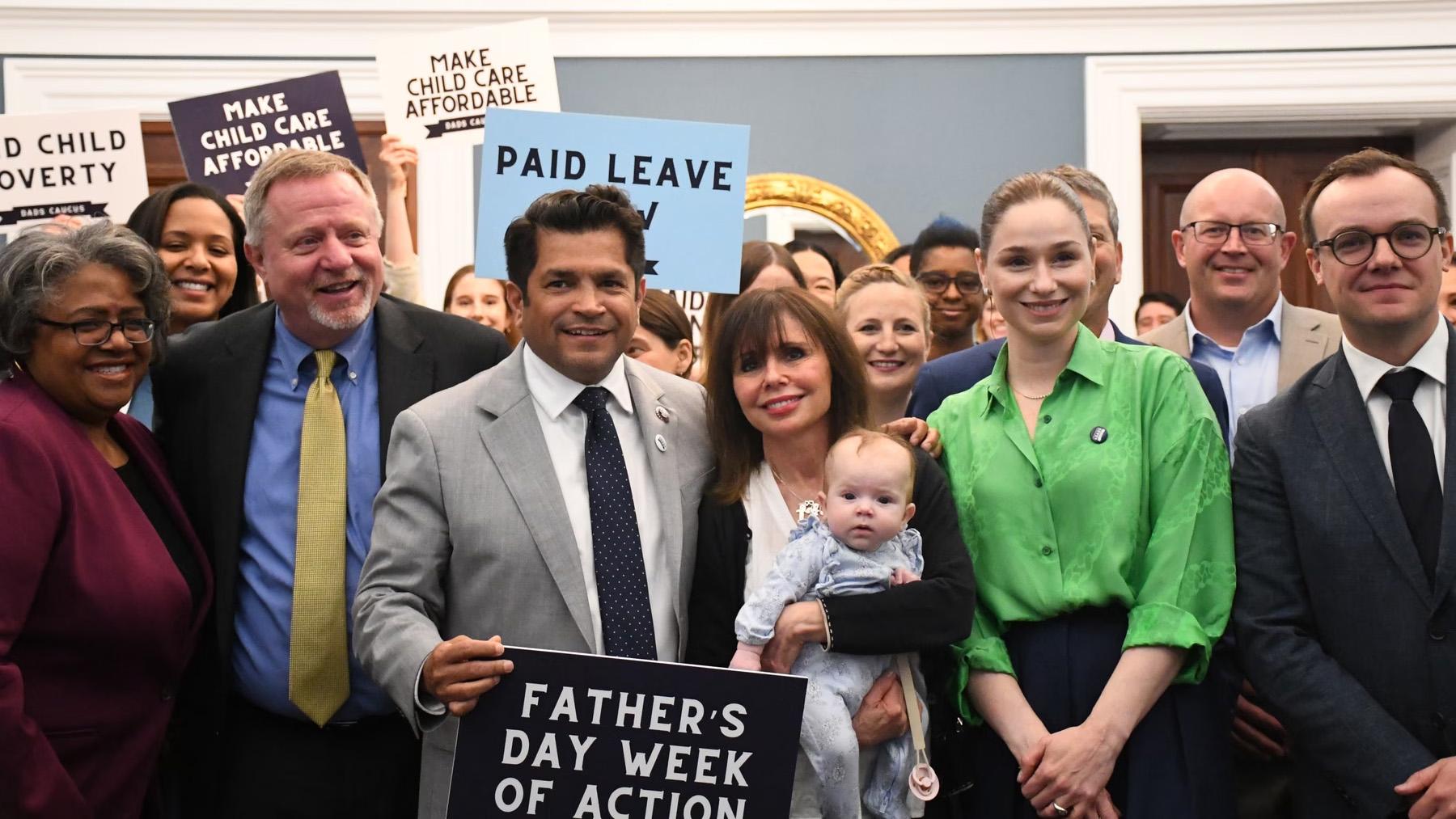 Group of people at an event holding signs about child care affordability and paid leave, with a woman holding a baby in the center.