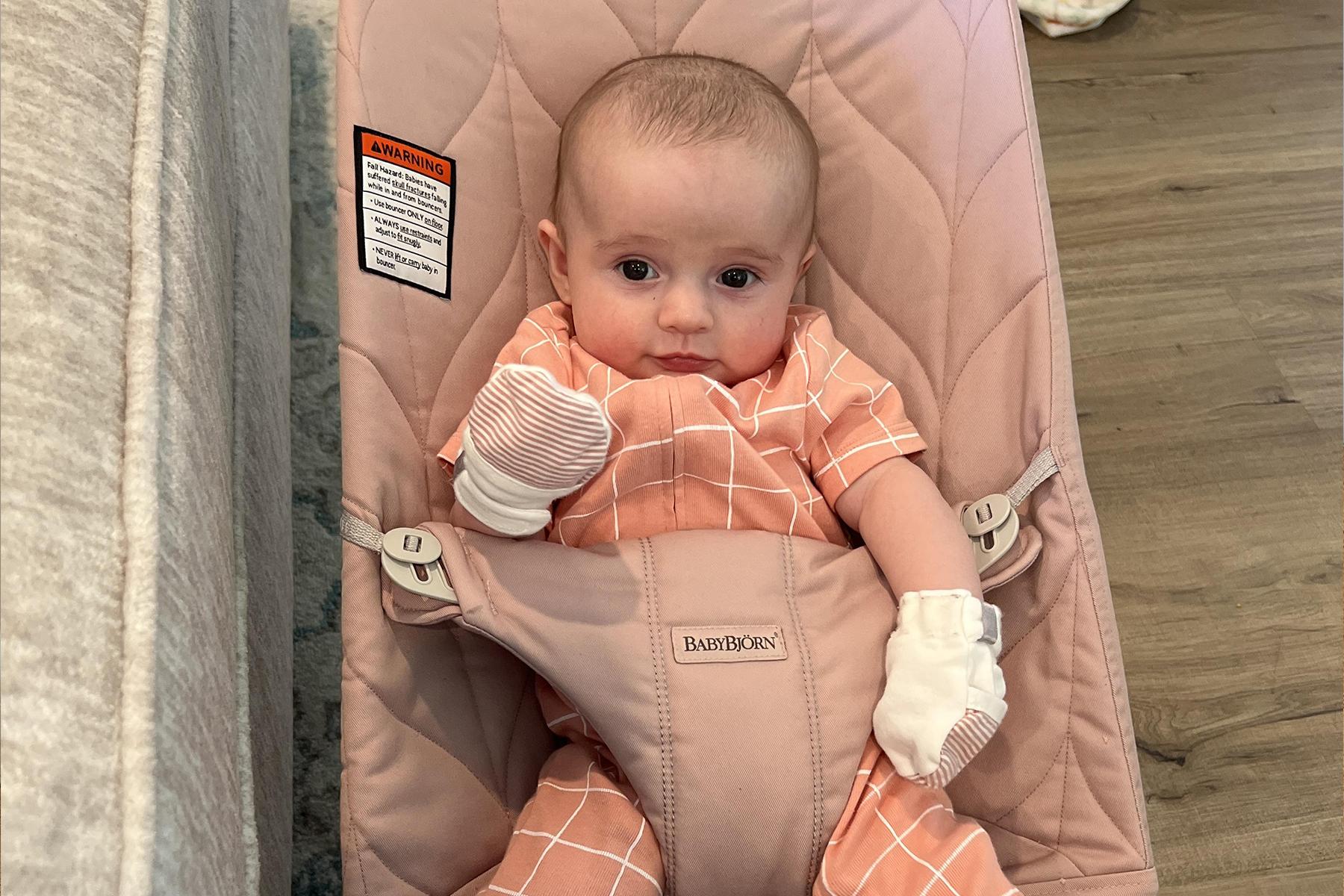 A baby in a pink bouncer wearing a peach outfit with white grid lines and mittens, sitting on a wooden floor.