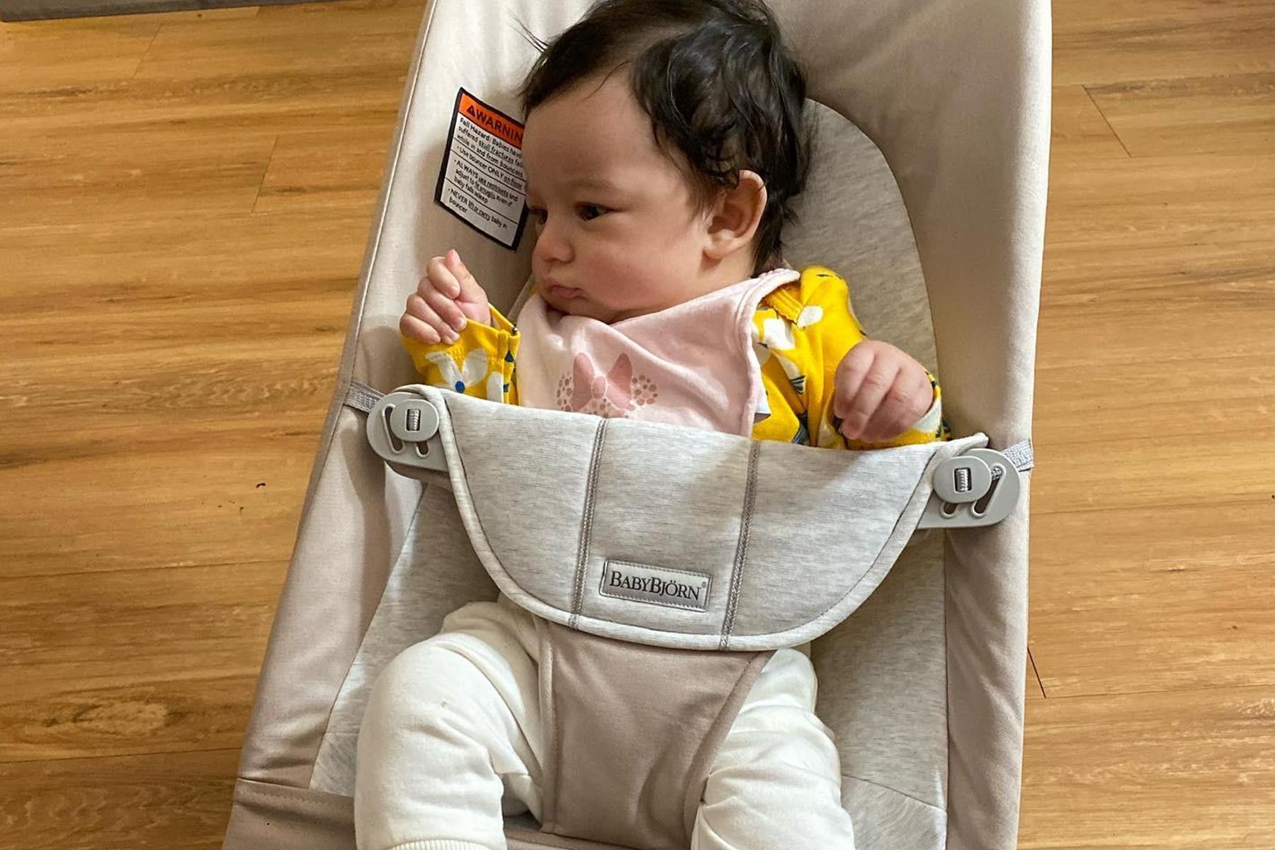 A baby in a yellow outfit sits in a beige baby bouncer on a wooden floor, looking to the side with a curious expression.