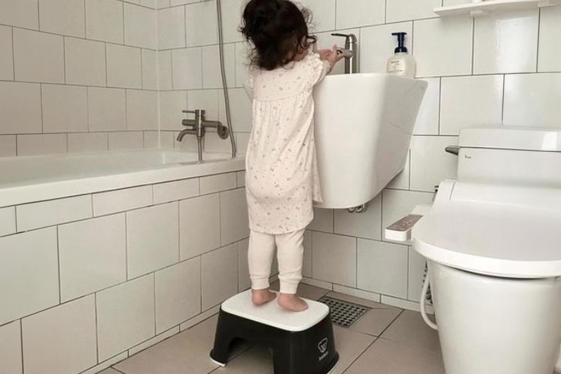 A young child stands on a small step stool at a bathroom sink, reaching for the tap. She wears pajamas in a tiled bathroom.