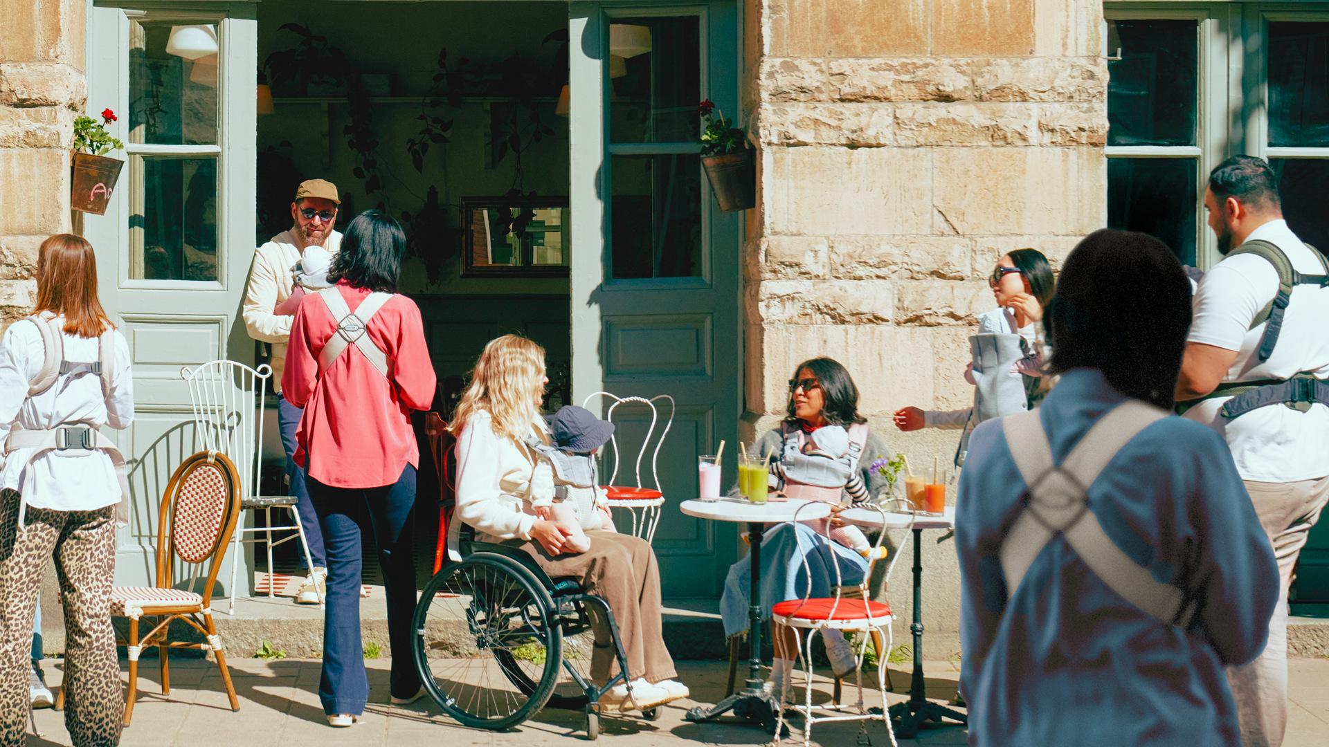 People socializing outside a café, with one person in a wheelchair and others sitting or standing, enjoying drinks and conversation in the sun.