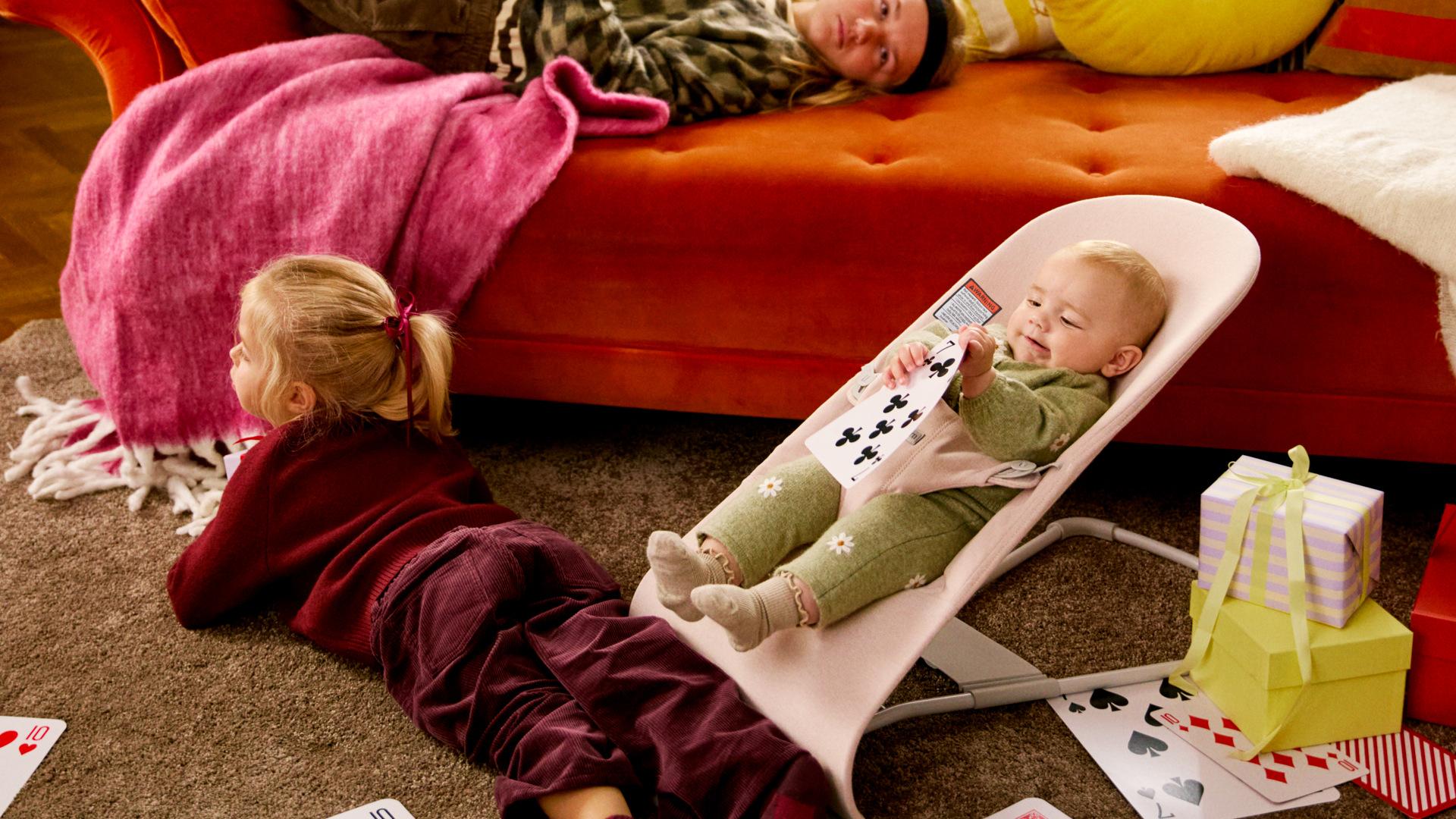 A baby in a bouncer holds playing cards, a child lies on the floor, and a person rests on an orange sofa with a pink blanket nearby.