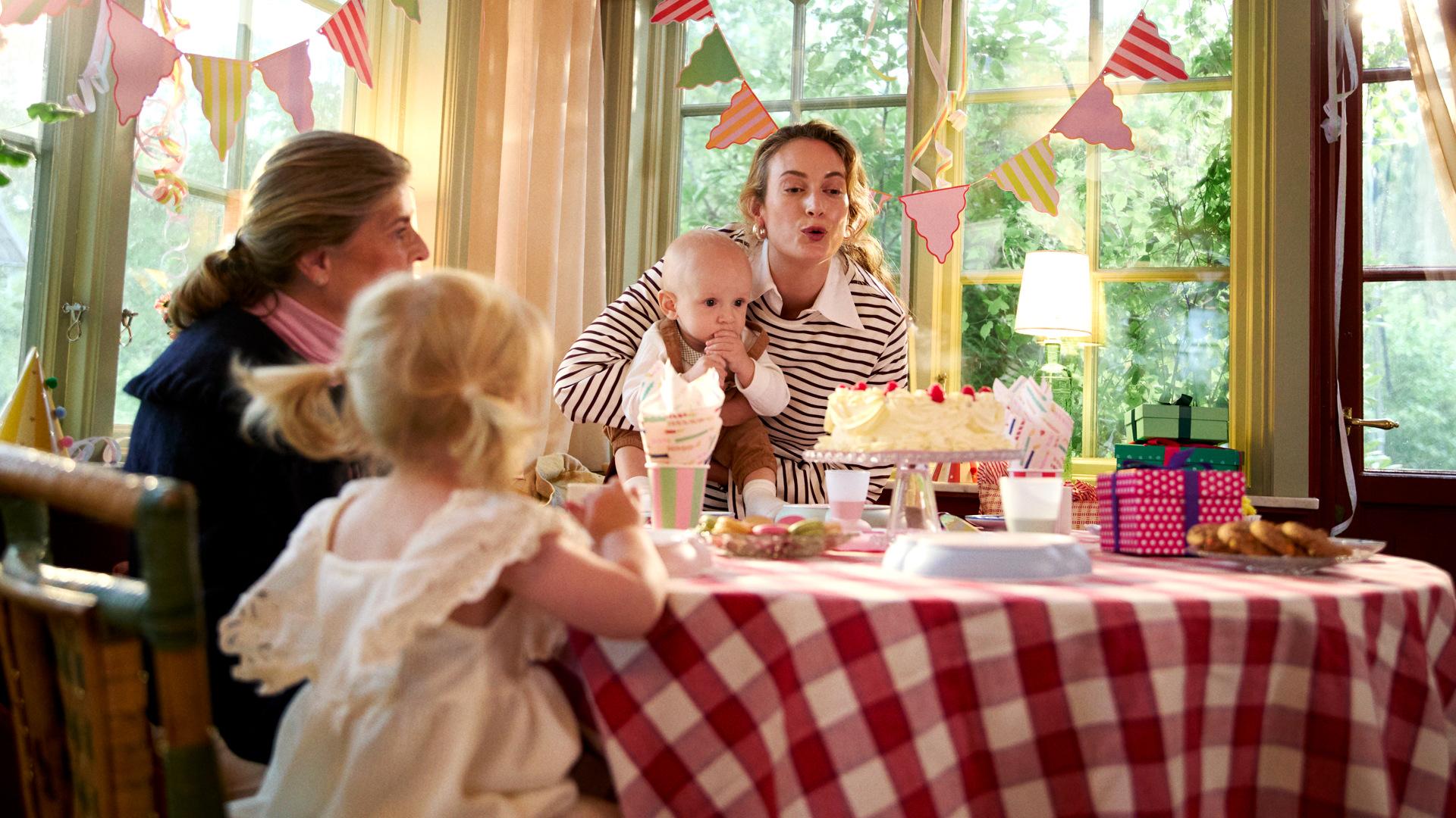 A woman holds a baby, blowing out candles on a cake at a festive table with children. Colorful bunting and gifts are in the background.