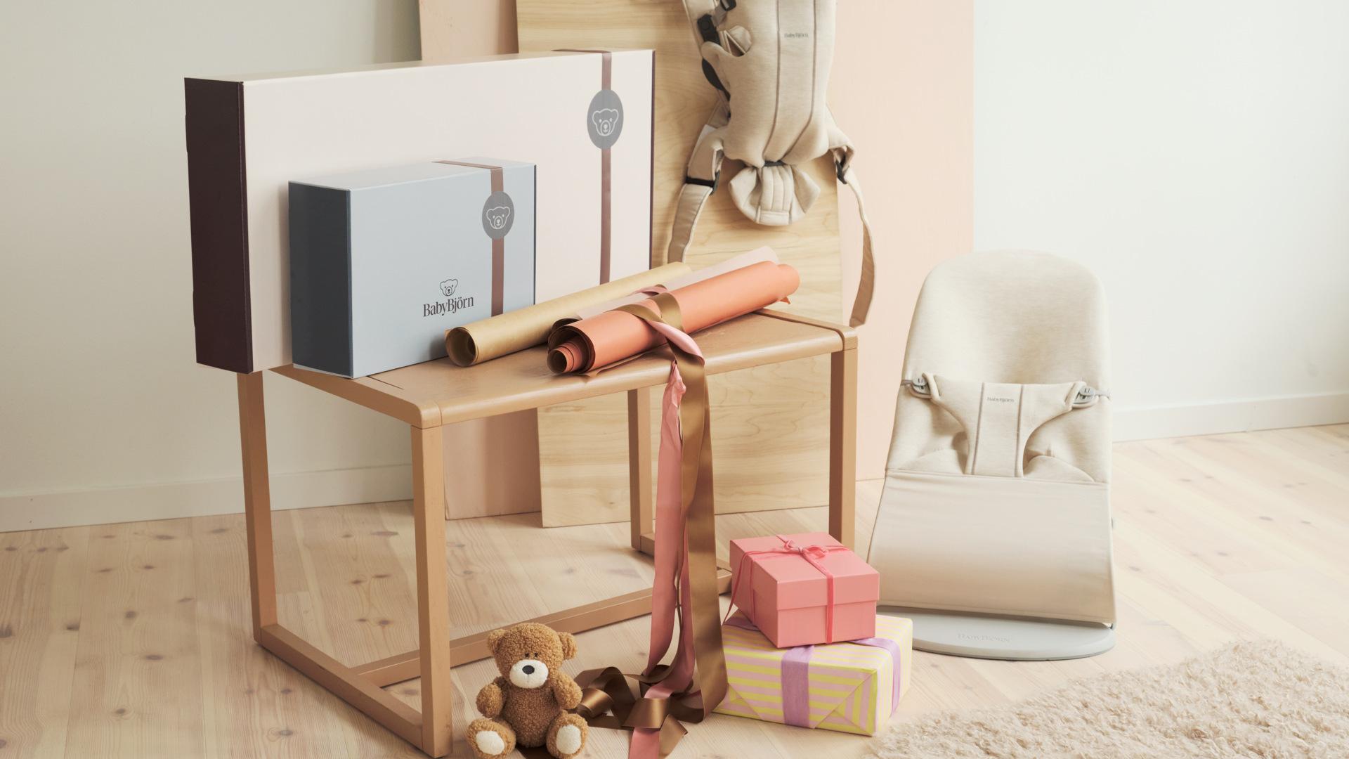 Nursery scene: gift boxes, rolled wrapping paper and ribbon on a wooden stool, teddy bears on floor beside a baby bouncer and backpack.