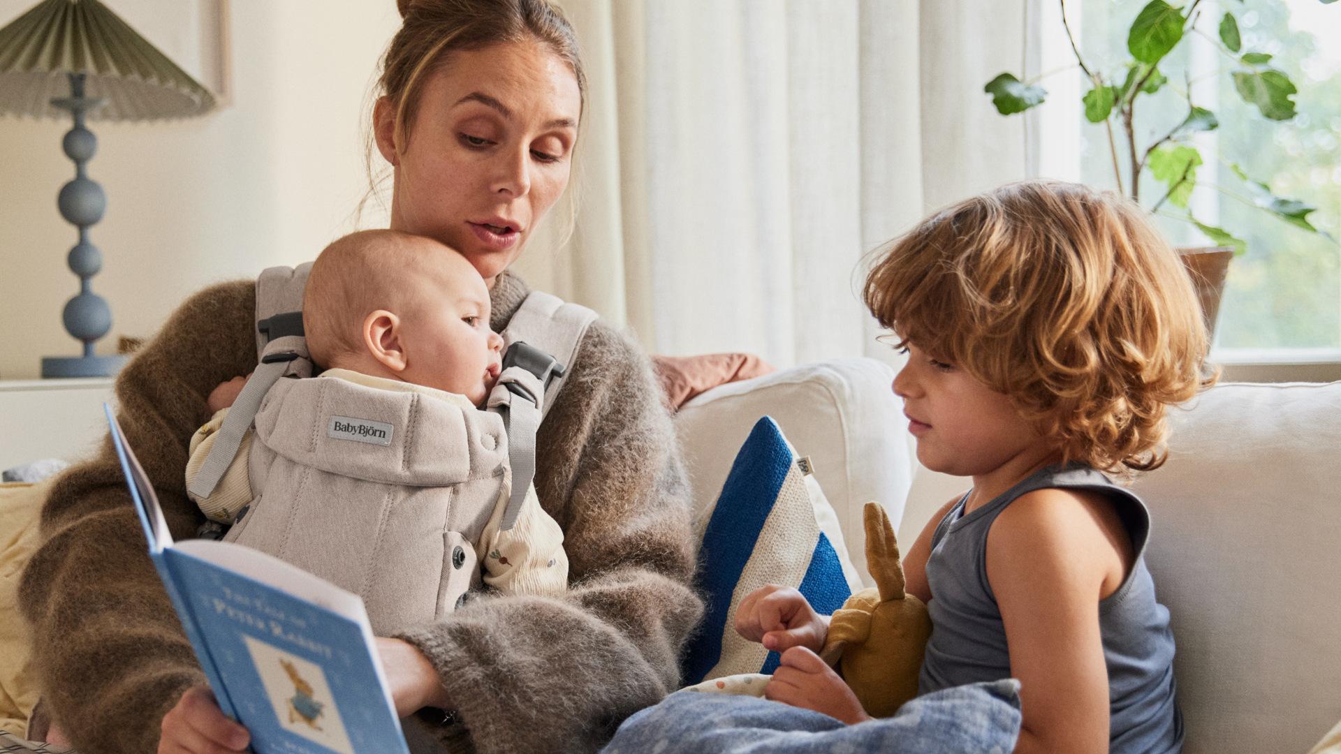 A woman reads a book to a baby in a carrier and a young child holding a stuffed toy, sitting on a couch in a cozy living room.