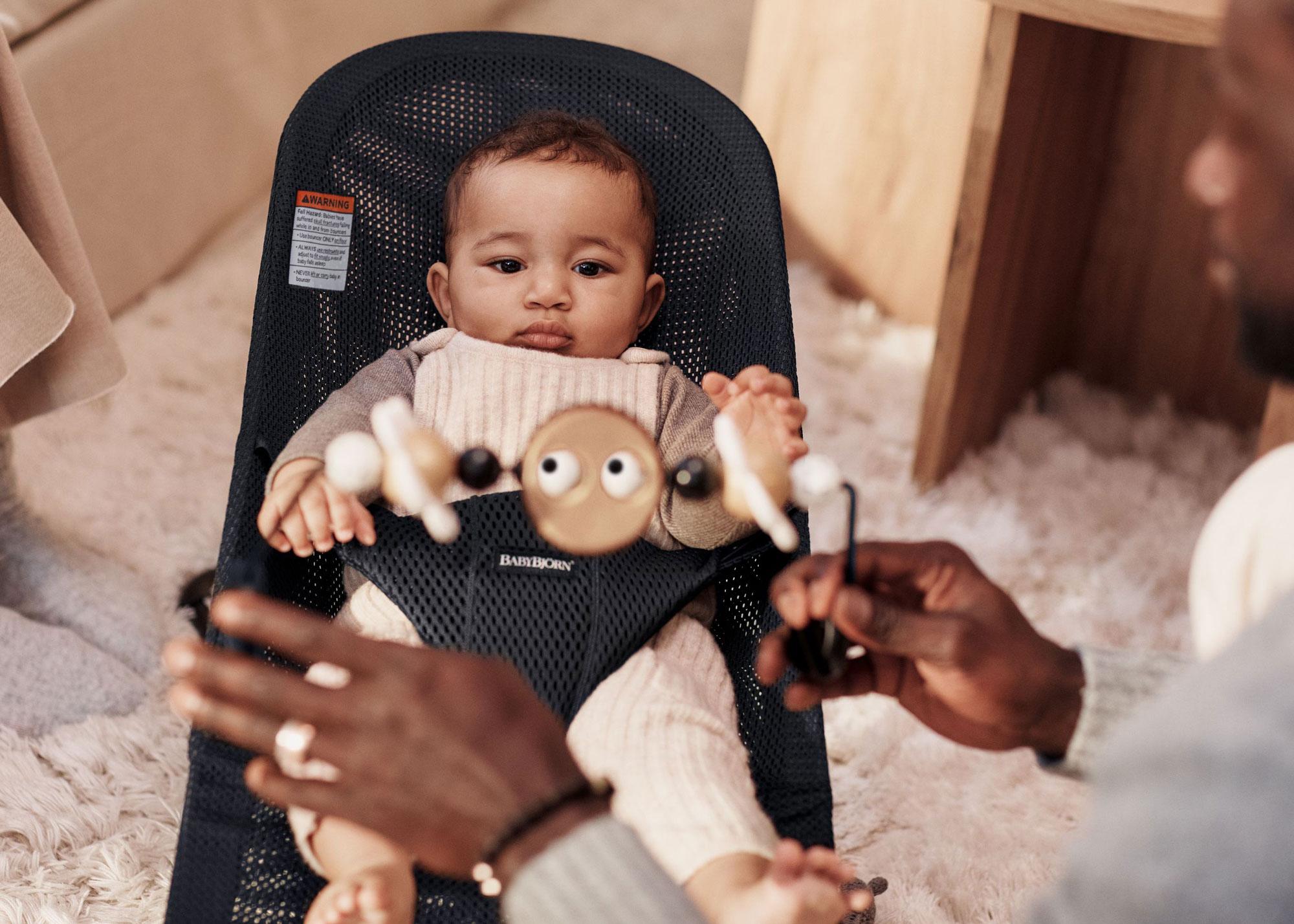 A baby sits in a bouncer, looking at a toy held by an adult. The room has a soft, cozy atmosphere with a fluffy rug.