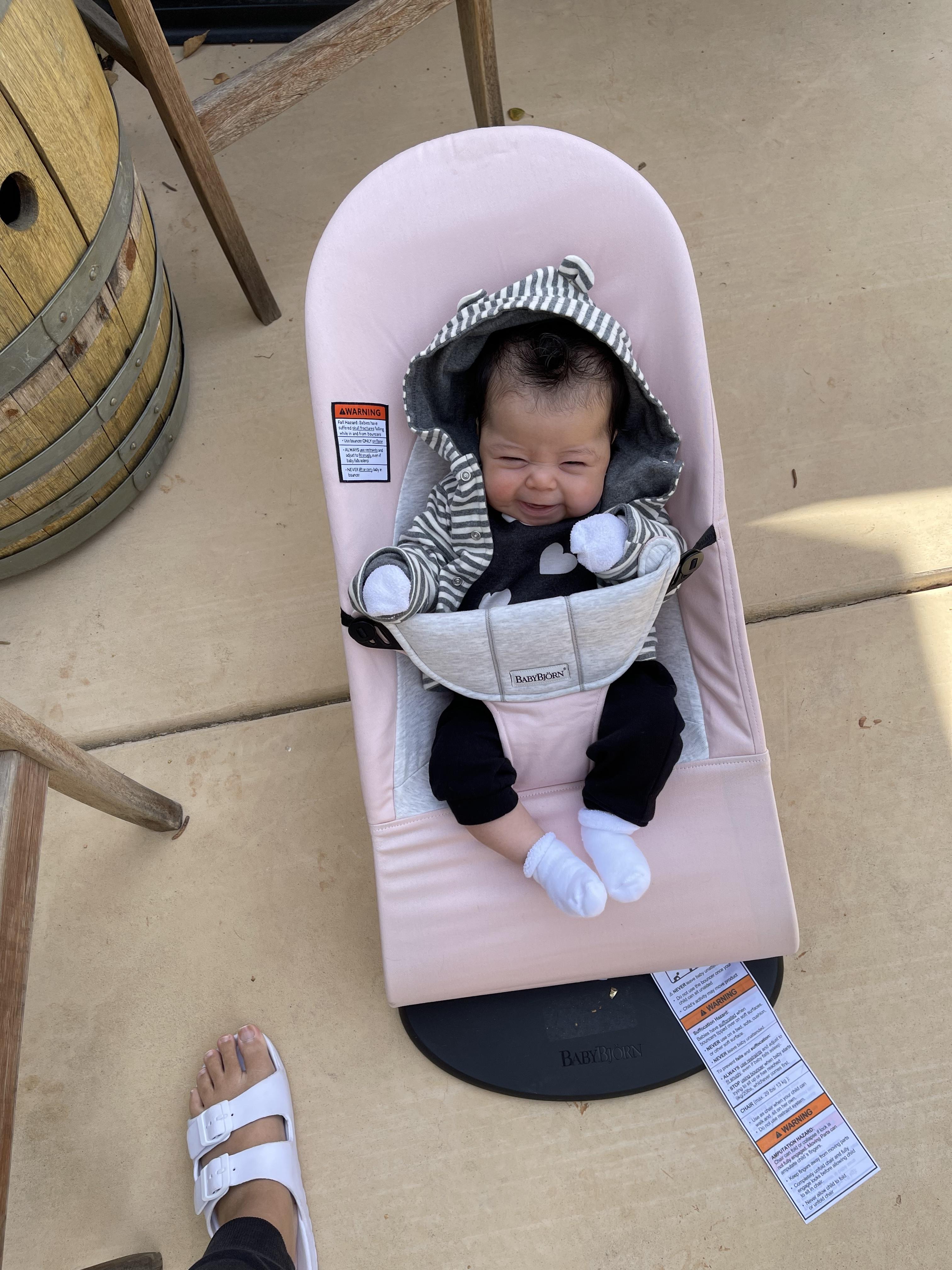 A smiling baby in a gray and white striped hoodie sits in a pink baby bouncer, with a wooden barrel and sandal in the background.